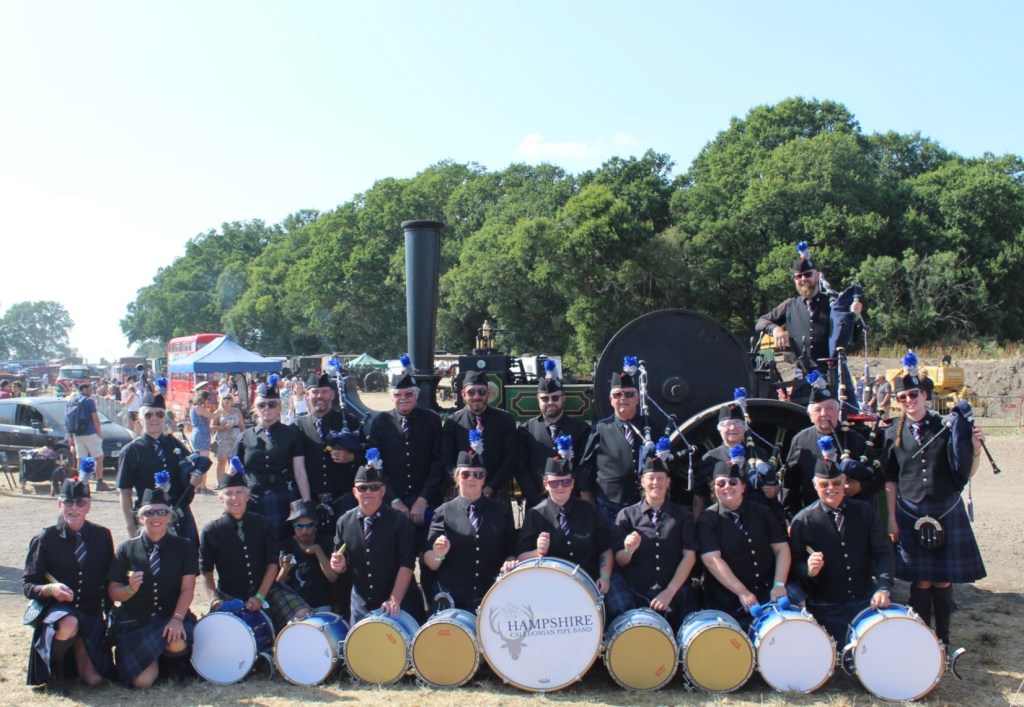 A pipe band having a group photo taken in front of a steam engine following a performance. The band are lined in rows with their bagpipes, and their drums lined up in front of the band.