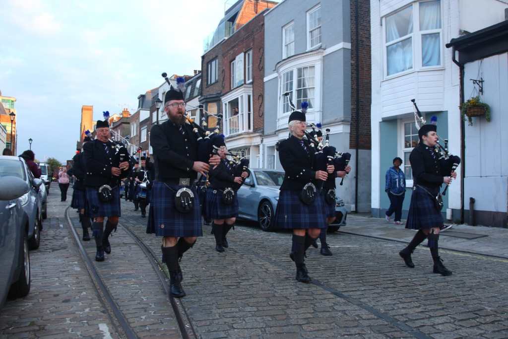 A pipe band marching down a street in their highland uniform and kilts, playing their bagpipes and drums.